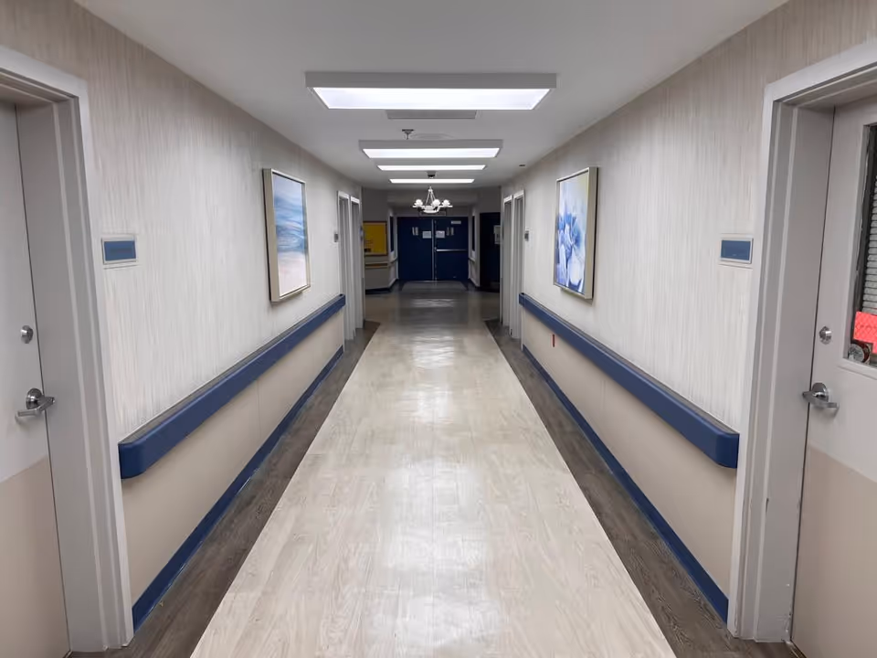 Long, well-lit interior hallway with handrails, doors, and framed artwork in a care facility.