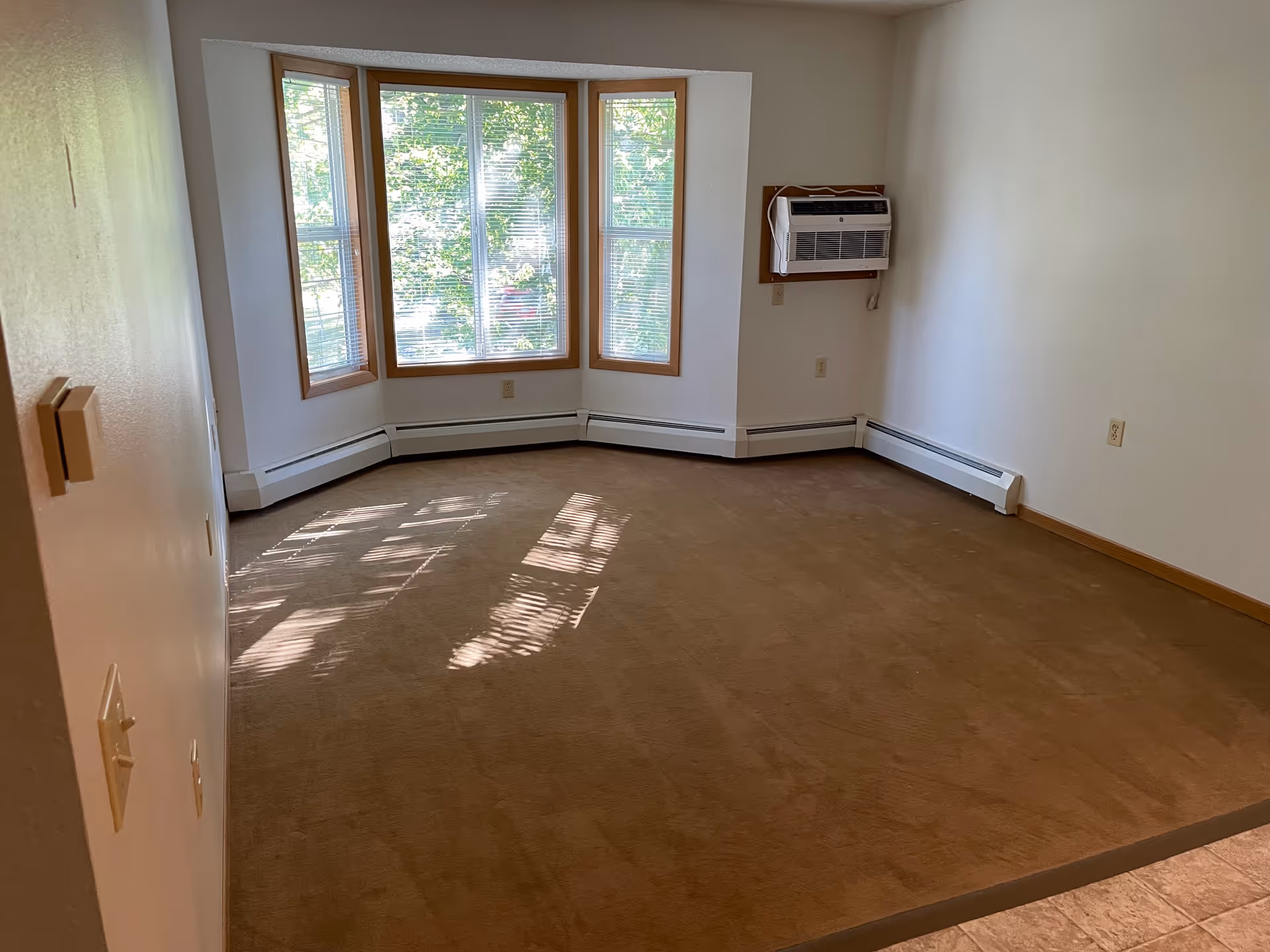 Empty room with beige carpet, white walls, a bay window with blinds, and a wall-mounted air conditioning unit.