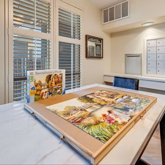 A table with a partially completed Ravensburger 500-piece puzzle depicting a scenic watermill and garden scene. Behind the table are windows with white blinds, a bulletin board labeled 'Gold Country Retirement Center,' and a wall with mailboxes and a letter slot.