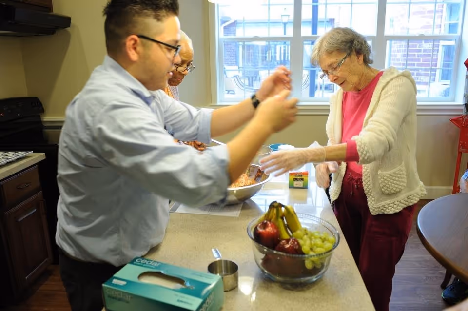 A man and two elderly women are preparing food together in a kitchen. The man is wearing glasses and a light blue shirt, while one elderly woman is wearing a white cardigan and pink shirt, and the other elderly woman is partially visible. They are standing around a kitchen island with a bowl of fruit and a large mixing bowl.