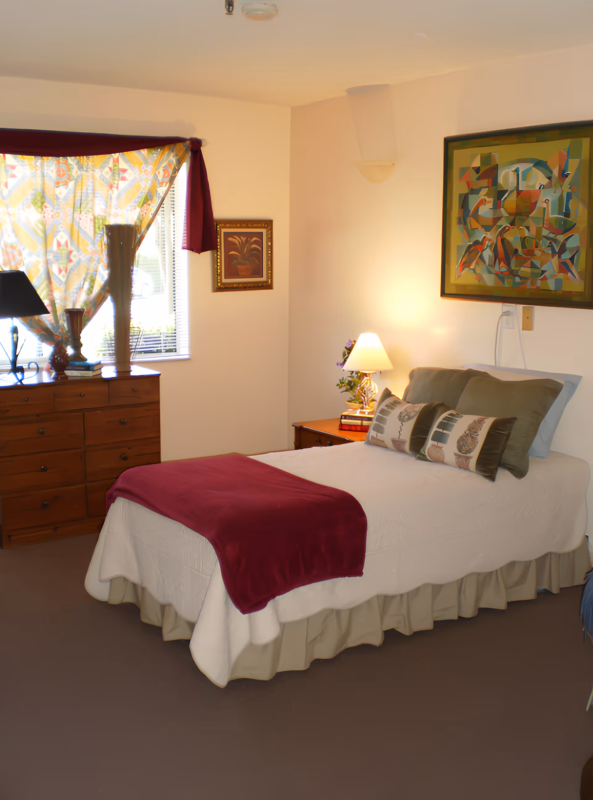 A cozy bedroom in Creekwood Care Facility featuring a neatly made bed with a white bedspread, green pillows, and a red throw blanket. There is a wooden dresser with decorative items and a window with colorful curtains. A bedside table with a lamp and a small plant is next to the bed. Two framed artworks hang on the walls.