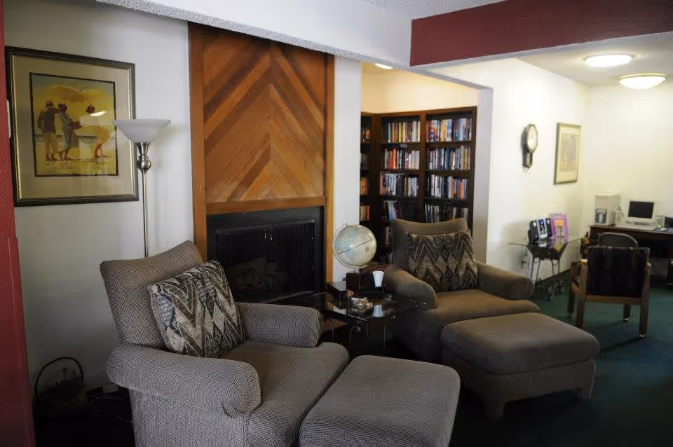 A cozy living room area with two cushioned armchairs and matching ottomans facing a wooden fireplace. There is a floor lamp and framed artwork on the wall to the left. In the background, there is a bookshelf filled with books and a desk with a computer and chair. The room has a carpeted floor and soft lighting.