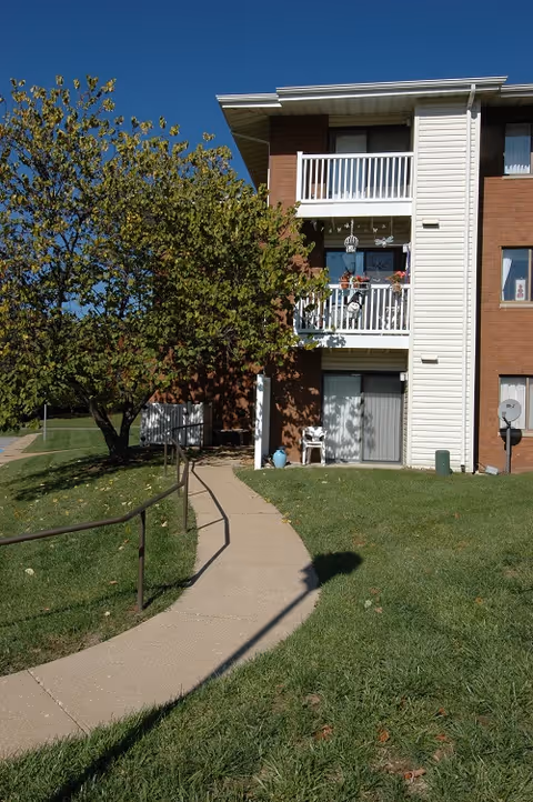 Exterior view of a three-story brick and siding building with balconies, a concrete pathway with a handrail leading to the entrance, and a tree with green leaves on a sunny day.