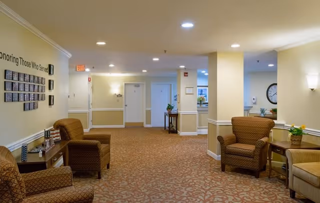 A well-lit hallway in an assisted living facility with patterned carpet, beige walls, and several comfortable armchairs arranged along the sides. There are small tables with decorative items and plants, wall sconces providing light, and a wall display honoring those who served. Doors and an exit sign are visible at the end of the hallway.