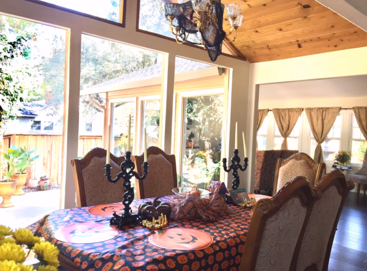 A dining room with a wooden table covered in a Halloween-themed tablecloth featuring pumpkin faces. The table is decorated with black candelabras holding white candles, small pumpkins, and other Halloween decorations. The room has large windows letting in natural light, with a view of an outdoor area with potted plants. The ceiling is wooden with a chandelier draped in black netting. There are upholstered dining chairs around the table and curtains on the windows in the background.