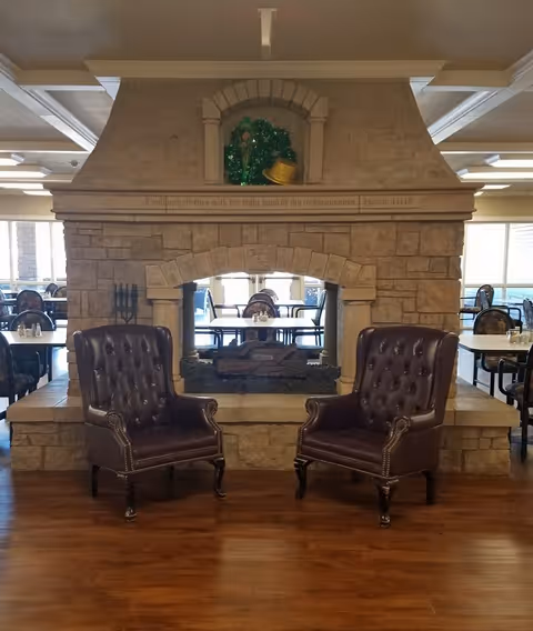 Two dark brown leather armchairs with tufted backs and nailhead trim are positioned in front of a large stone double-sided fireplace in a spacious room with wooden floors and multiple dining tables and chairs visible in the background.