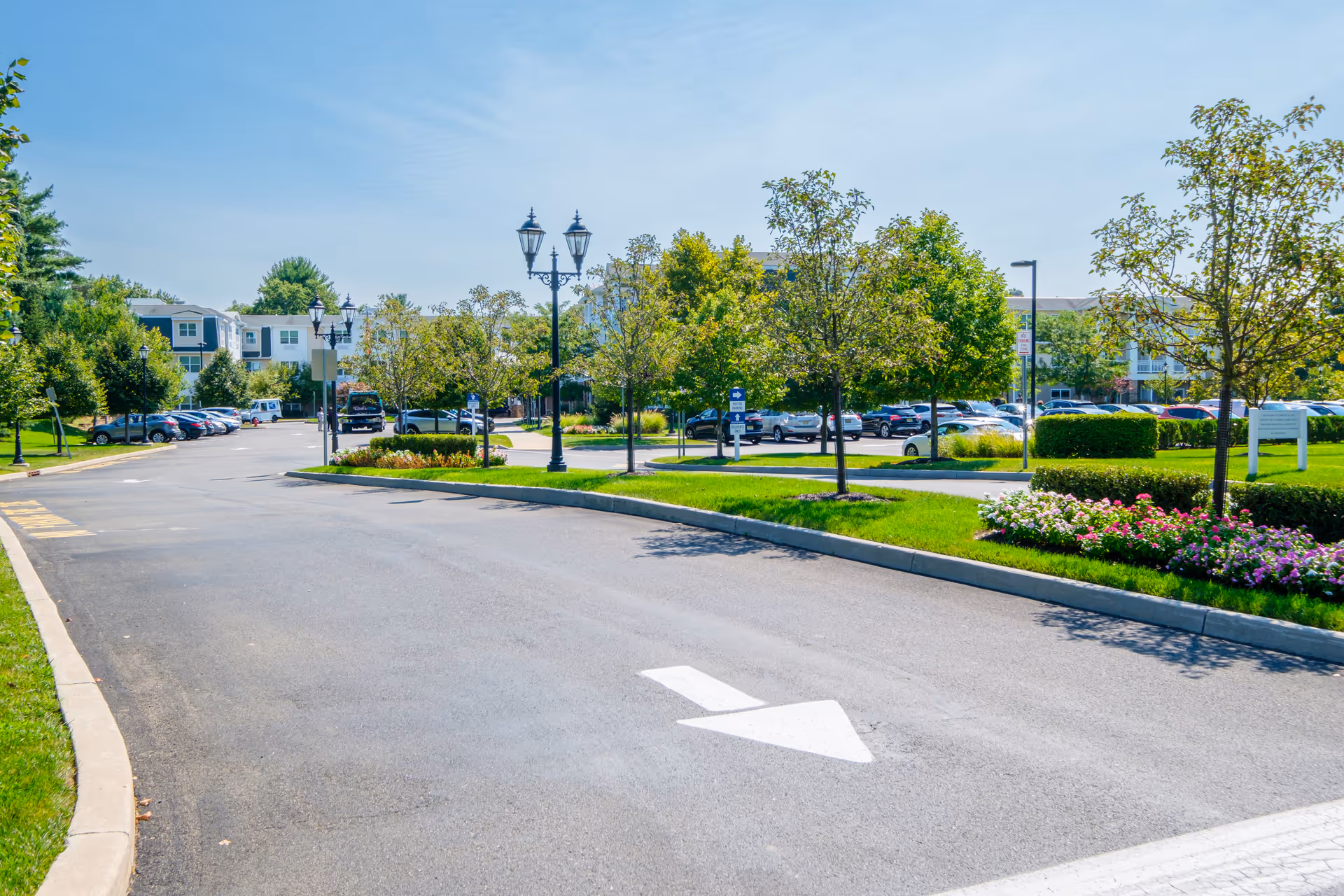 A paved driveway with a white directional arrow pointing forward, flanked by green grass, trees, and flower beds. There are parked cars and a multi-story building in the background under a clear blue sky.