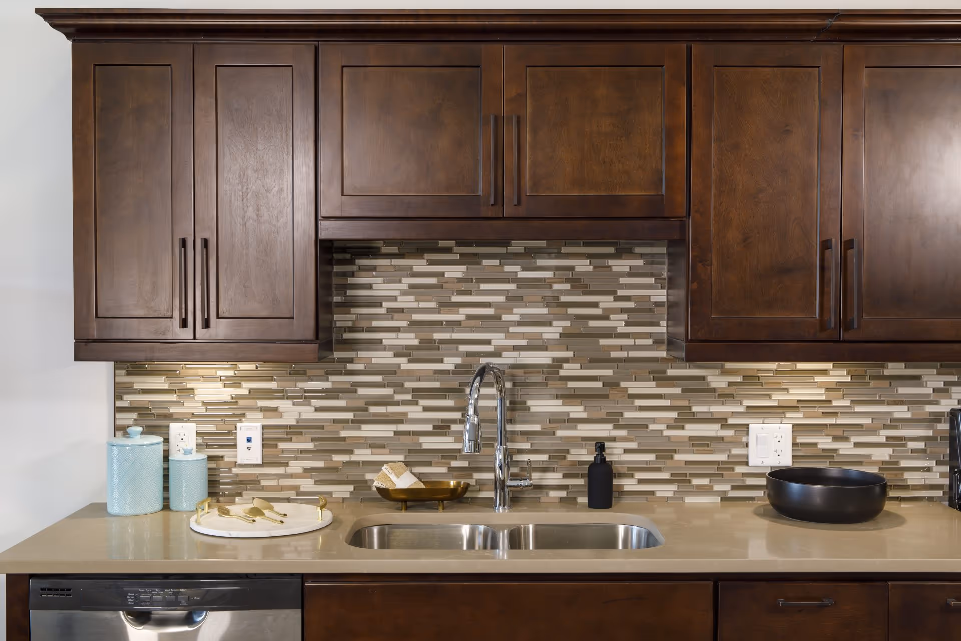Modern kitchen area with dark wooden cabinets, a beige countertop, a stainless steel sink with a chrome faucet, and a mosaic tile backsplash in shades of beige, brown, and cream. On the countertop are two light blue canisters, a round tray with wooden utensils, a soap dispenser, and a black bowl. A dishwasher is visible below the counter.