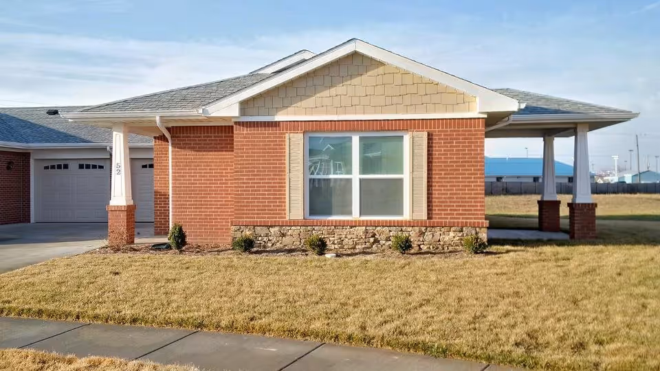 Front exterior view of a single-story brick building with a large window, beige shutters, and a covered porch area on the right side. The building has a shingled roof and a small landscaped area with grass and shrubs in front.