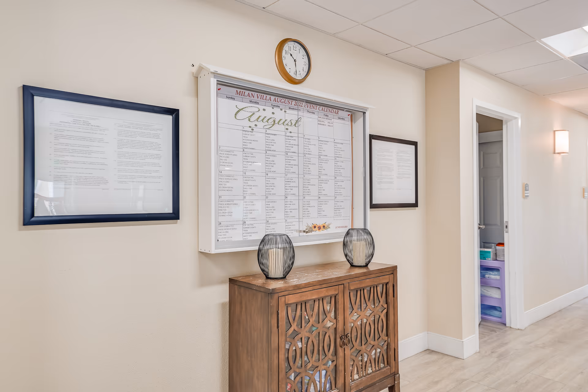 Hallway interior with a wall-mounted event calendar and clock above a wooden cabinet holding two decorative vases.