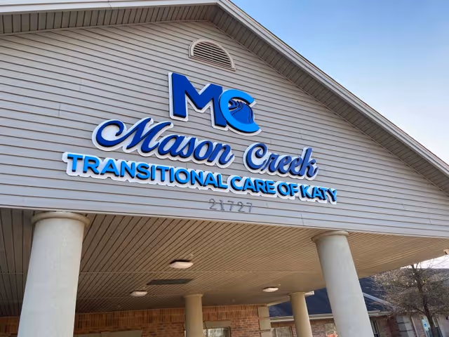 Exterior view of the entrance to Mason Creek Transitional Care of Katy facility, showing the building's name and logo on the facade above the covered entry supported by columns.
