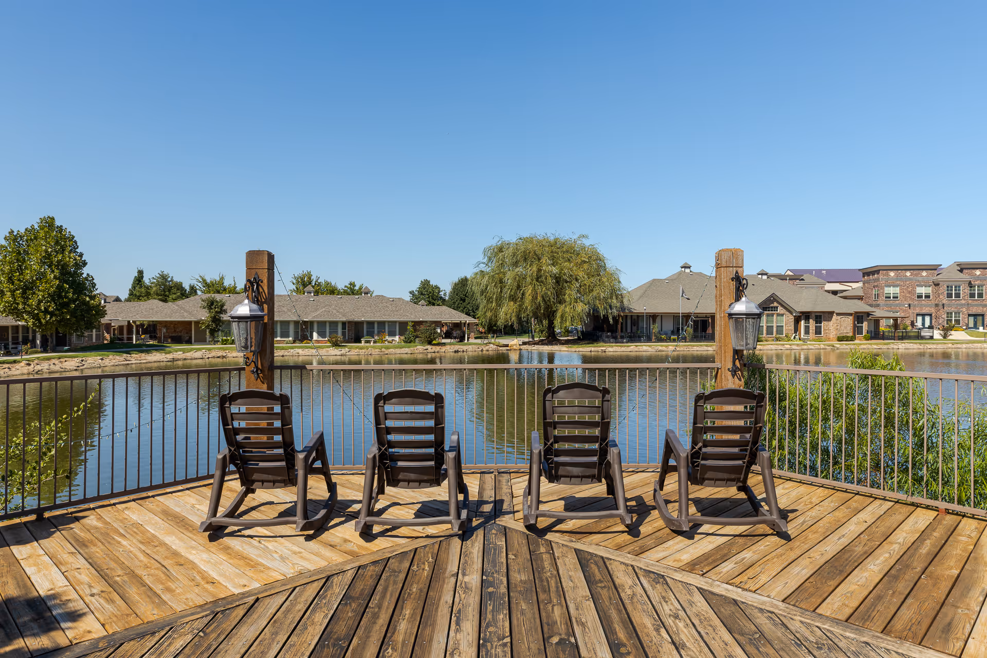 A wooden deck with four brown rocking chairs facing a calm pond. The deck has metal railings and two wooden posts with lantern-style lights. Across the pond, there are single-story and multi-story buildings surrounded by trees and greenery under a clear blue sky.