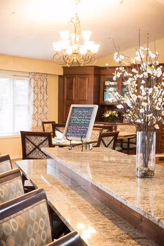 A bright dining area with a large granite countertop island surrounded by cushioned chairs with patterned upholstery. In the background, there is a wooden cabinet with glass doors and a window with patterned curtains. A chandelier with multiple lights hangs from the ceiling, and a tall vase with white flowers is placed on the countertop. A small chalkboard menu is displayed on the island.