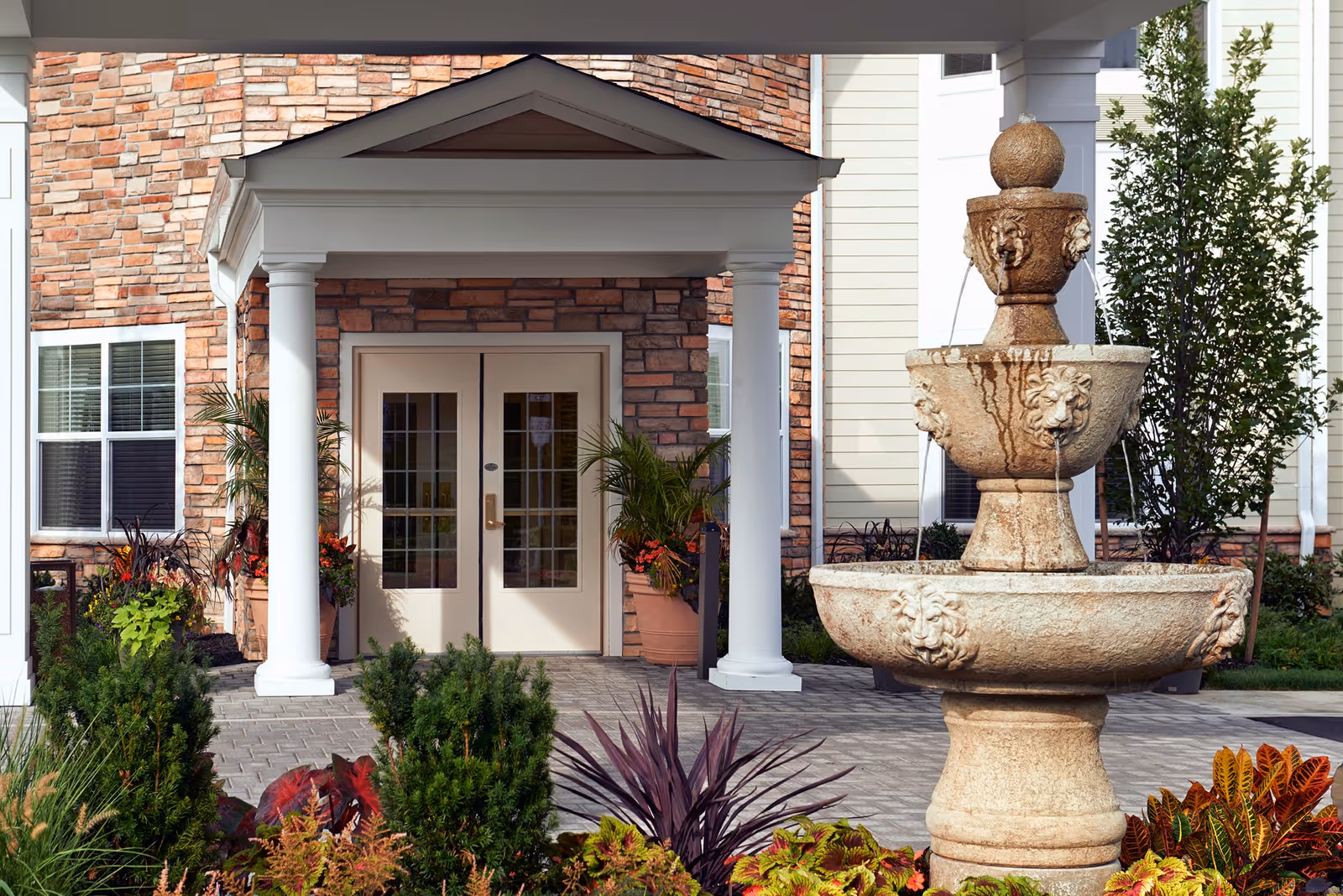 Entrance to a building with double glass doors framed by white columns and a small roof. In front of the entrance is a tiered stone fountain with water flowing, surrounded by various plants and shrubs. The building exterior features a mix of stone and siding.