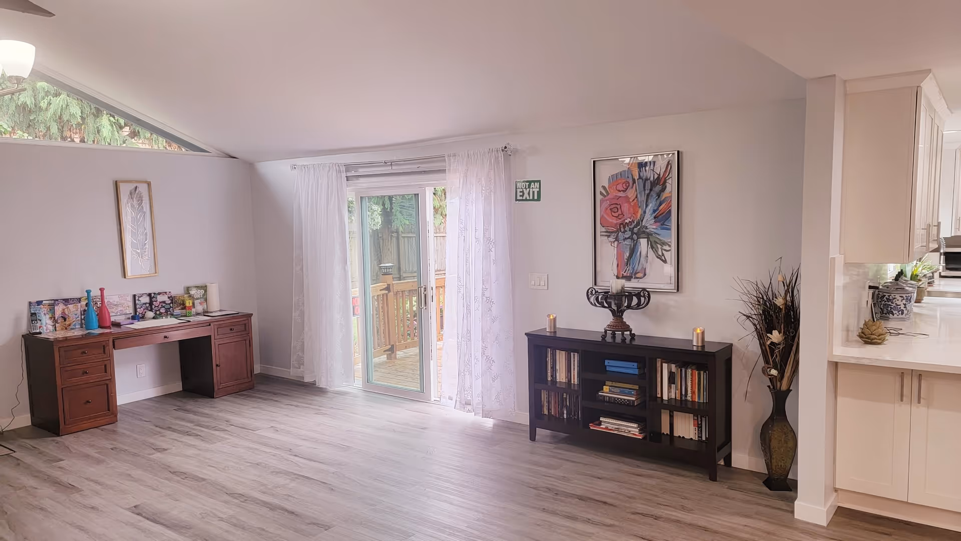 Open living room with light wood floors, a desk on the left, sliding glass doors to a deck, and a bookshelf with artwork on the right.
