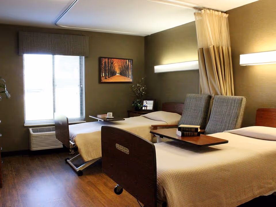 Two hospital-style beds with overbed tables and chairs in a well-lit senior care room with a window and wall art.