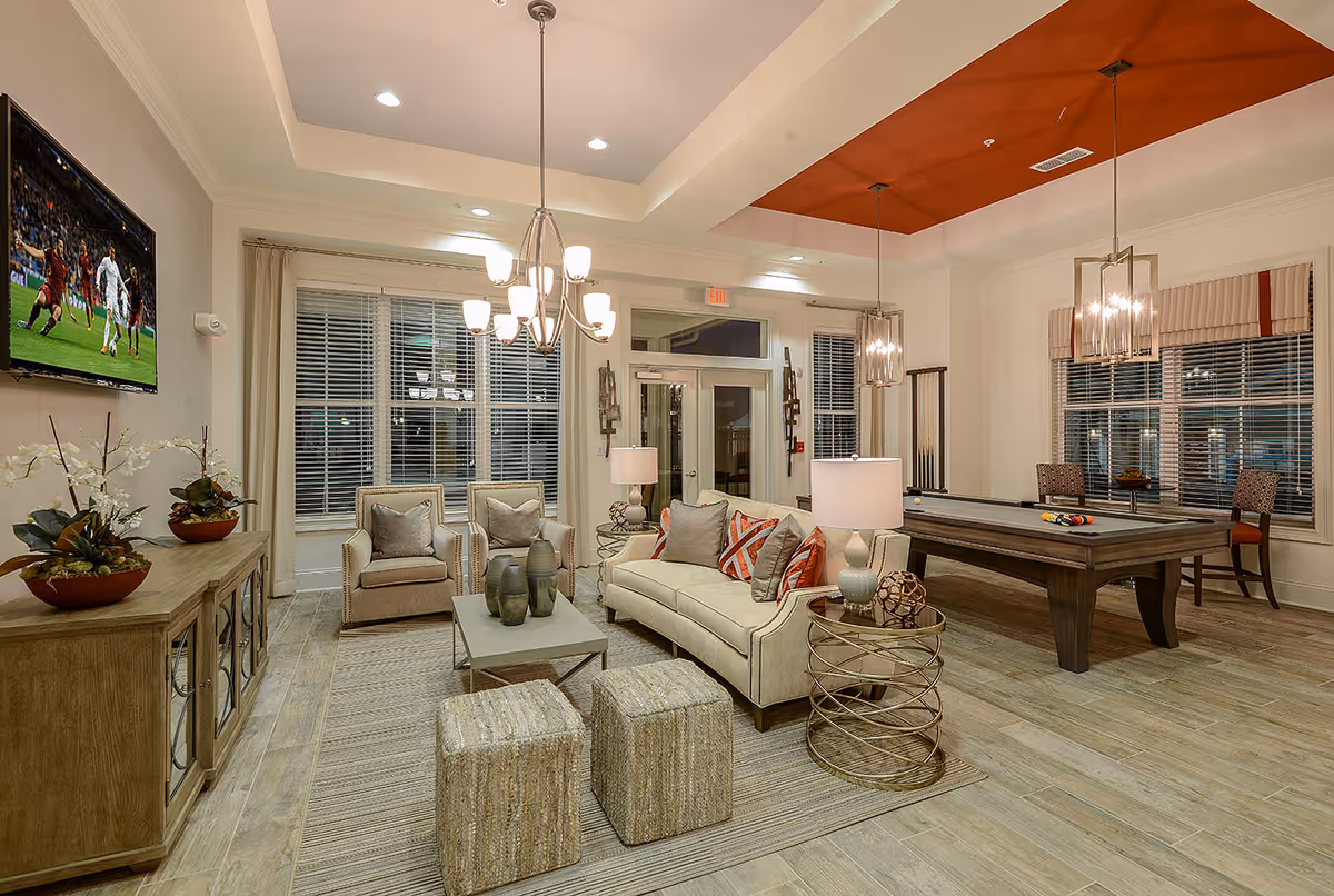 Well-lit communal living room/lounge with sofas, chairs, a coffee table, wall-mounted TV and a pool table under pendant lights.