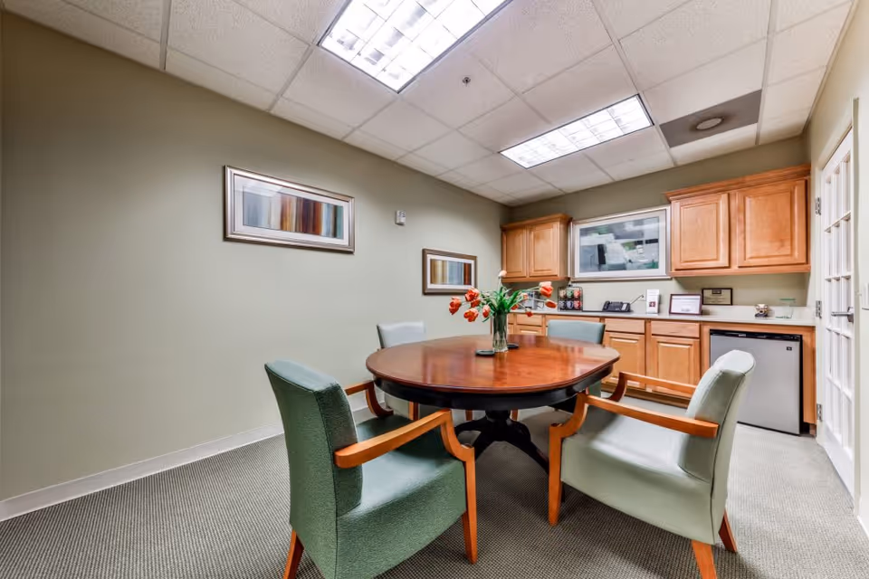 A small meeting or dining room with a round wooden table surrounded by four green upholstered chairs with wooden armrests. On the table is a vase with orange tulips. The room has light green walls with two framed abstract paintings. There are wooden cabinets and a small refrigerator along the back wall under fluorescent ceiling lights.