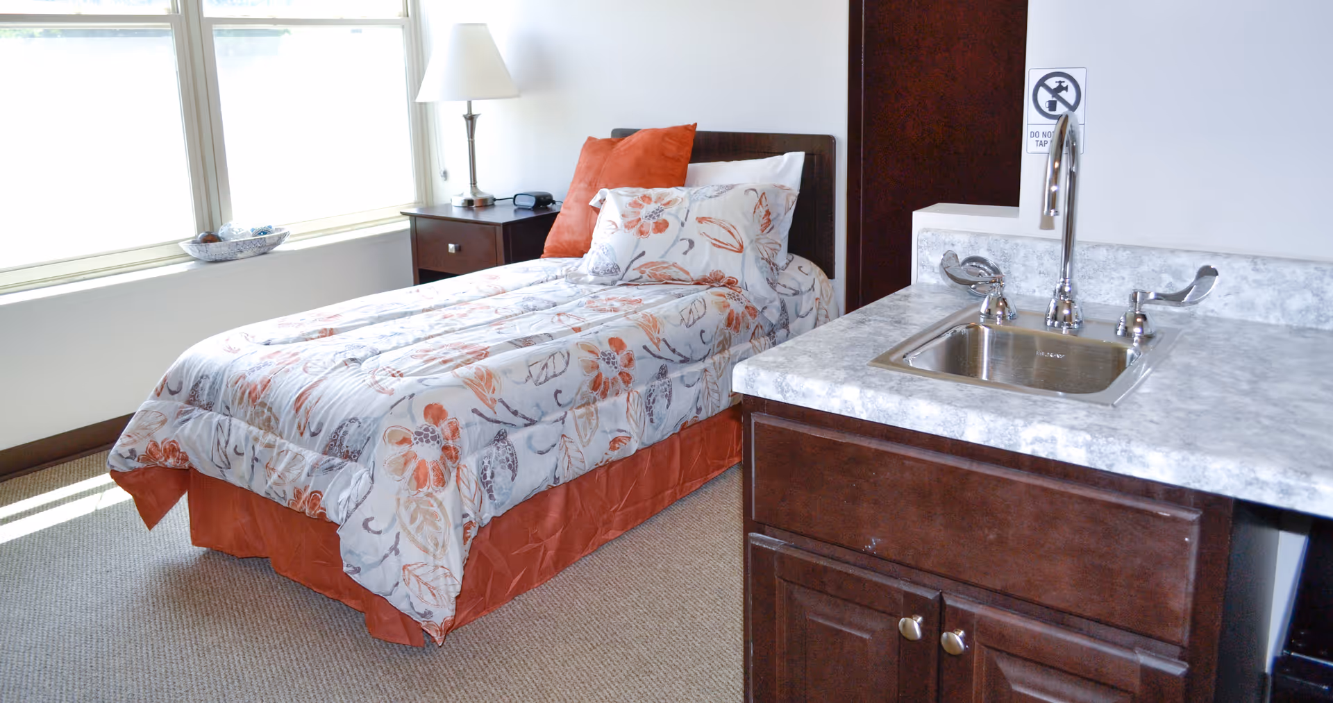 A single bed with floral bedding and orange accents is positioned next to a window with blinds. Beside the bed is a dark wooden nightstand with a lamp and a small decorative bowl on the windowsill. To the right, there is a countertop with a small sink and a faucet, along with a sign above the sink that says 'Do Not Tap'.
