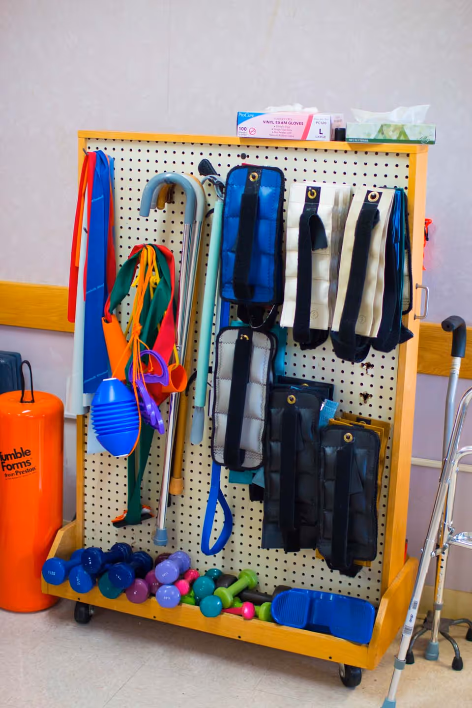 A wooden pegboard on wheels holding various physical therapy and exercise equipment including resistance bands, ankle weights, canes, and small dumbbells. There is a box of vinyl exam gloves and a box of tissues on top of the pegboard. An orange cylindrical object labeled 'Fumble Forms' is next to the pegboard, and a walker is partially visible on the right side.
