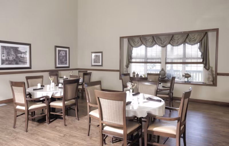 A dining room with several round tables covered with white tablecloths, each surrounded by wooden chairs with cushioned seats and backs. The room has light-colored walls adorned with framed artwork and a large window with blinds and decorative curtains, allowing natural light to enter.