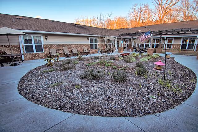 Circular mulched courtyard with seating, planters and an American flag surrounded by a single-story brick senior living building.