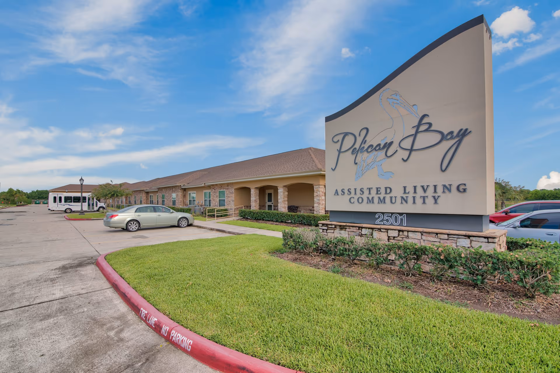 Exterior view of Pelican Bay Assisted Living Community building with a large sign displaying the facility name and address 2501. The building has a beige facade with stone accents and a covered walkway. Several cars are parked in the parking lot under a partly cloudy blue sky.