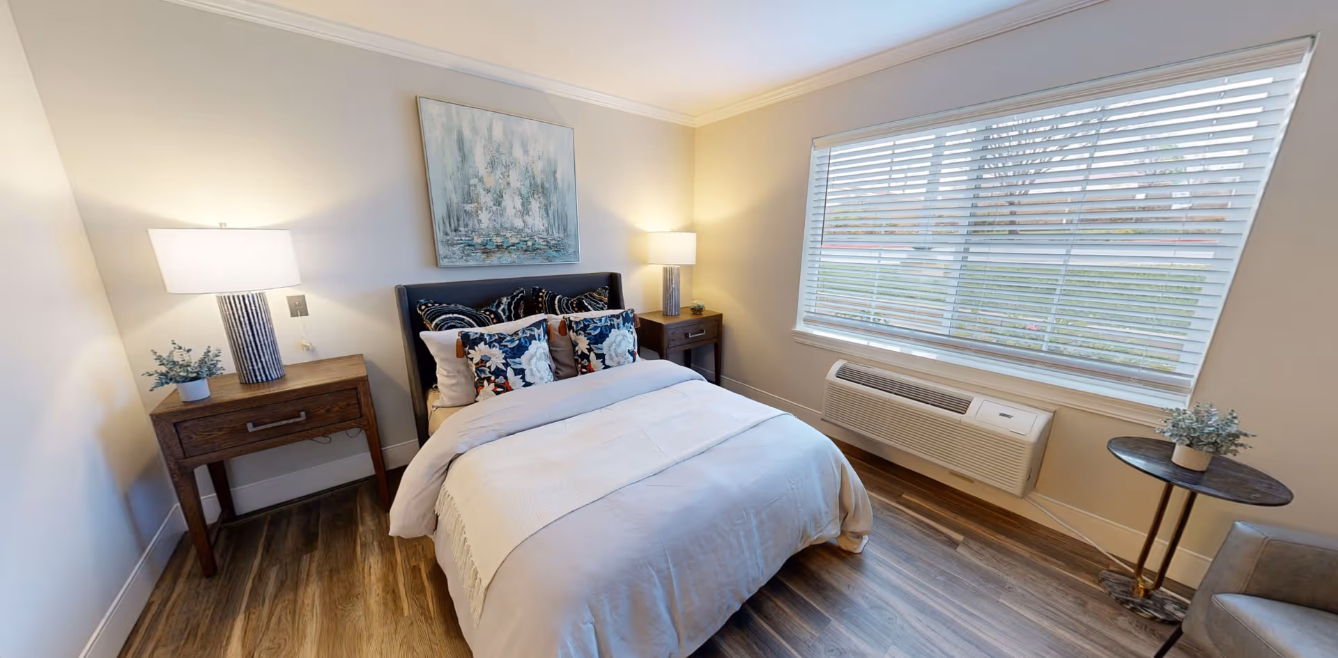 A cozy bedroom with a neatly made bed featuring multiple decorative pillows. There are two wooden nightstands on either side of the bed, each with a lamp and a small plant. A large window with blinds lets in natural light, and a piece of abstract artwork hangs above the bed. The room has wood flooring and neutral-colored walls.