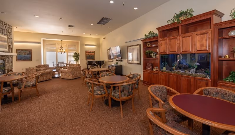 Community lounge and dining area with round tables and chairs, upholstered sofas, a fireplace, wall-mounted TV, and a large built-in aquarium.