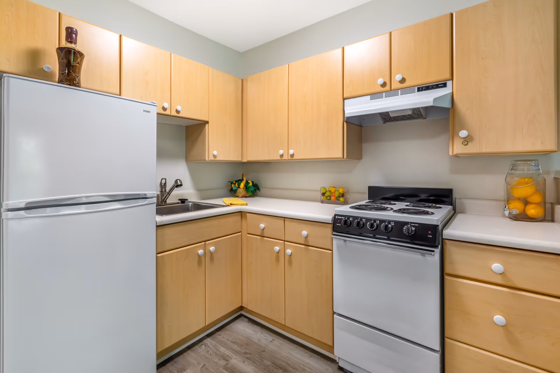 A compact kitchen with light wood cabinets, a white refrigerator, a white electric stove with an overhead vent, a stainless steel sink, and a light-colored countertop. There are decorative items including a basket with lemons, a glass container with oranges, and a small container with assorted fruit on the countertop.