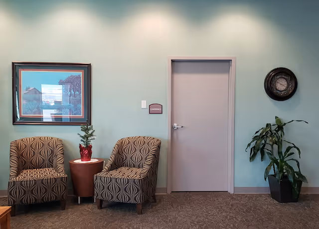 A waiting area with two patterned armchairs separated by a small round table holding a small potted plant. On the wall behind is a framed picture, a closed door with a sign labeled 'Conference', and a round wall clock. A tall potted plant is placed to the right of the door.