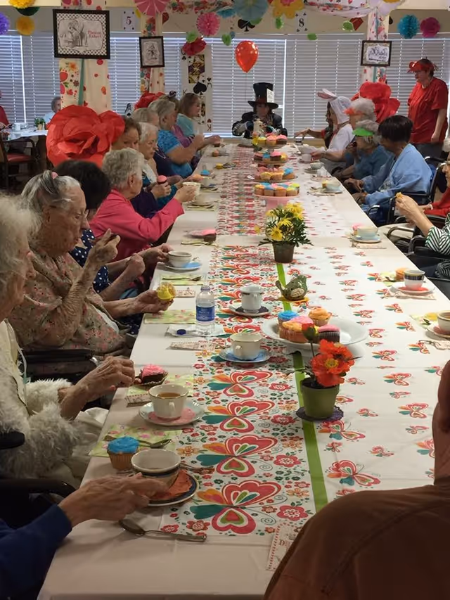 A group of elderly people seated around a long table decorated with colorful floral and heart-patterned table runners, enjoying cupcakes and tea. The room is decorated with large paper flowers, hanging paper decorations, and playing card-themed wall art. Some individuals are wearing festive hats and costumes, suggesting a themed party or celebration.