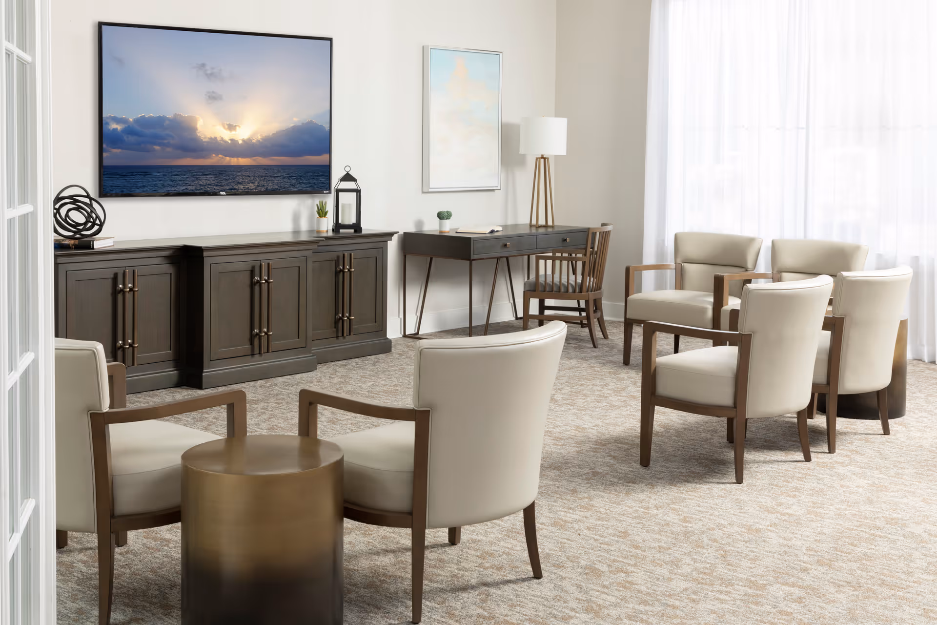 A bright and spacious living room with beige carpet and cream-colored armchairs arranged around a small round bronze table. Against the wall is a dark wooden cabinet with decorative items and a flat-screen TV displaying a sunset over the ocean. A wooden desk with a chair and a floor lamp is positioned near a large window with sheer white curtains, allowing natural light to fill the room.