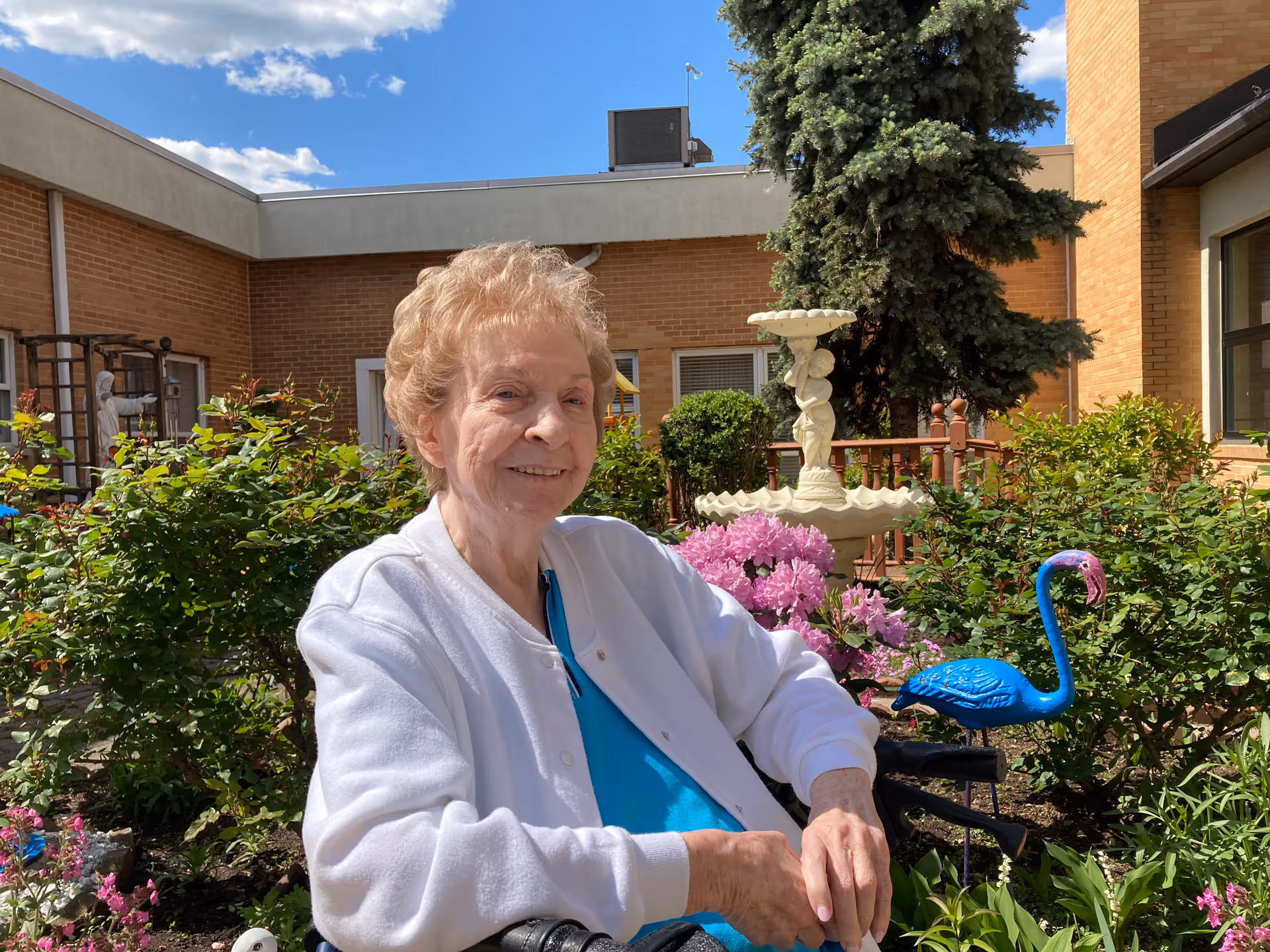 An elderly woman with short curly hair wearing a white cardigan and blue shirt sits in a wheelchair in a garden courtyard. Behind her are green bushes, pink flowers, a blue flamingo garden ornament, a white fountain, and a large evergreen tree. The courtyard is surrounded by brick buildings under a blue sky with some clouds.