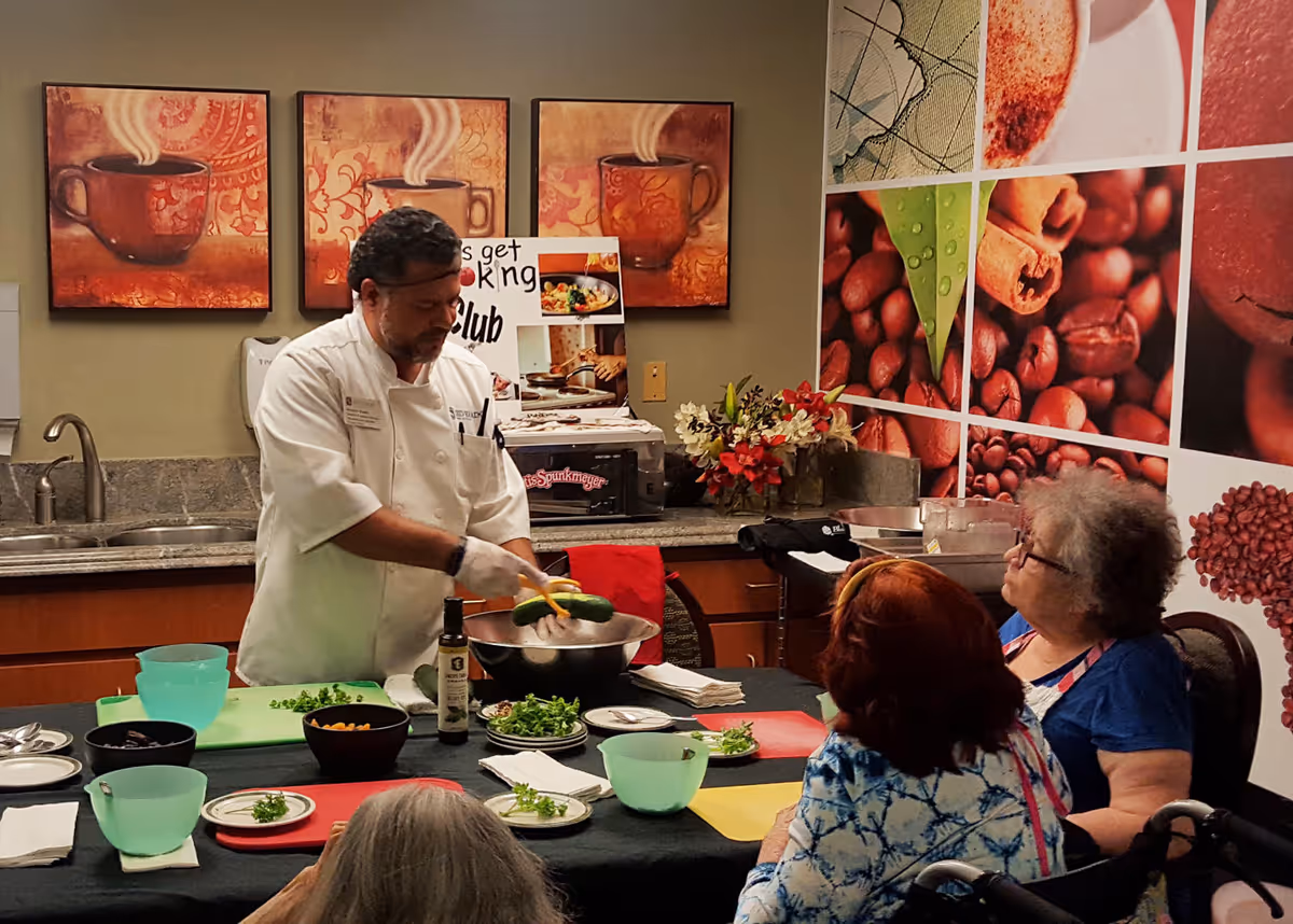 A chef demonstrating cooking techniques to a group of elderly women seated around a table in a kitchen area decorated with coffee-themed artwork and a large photo collage of coffee beans on the wall.