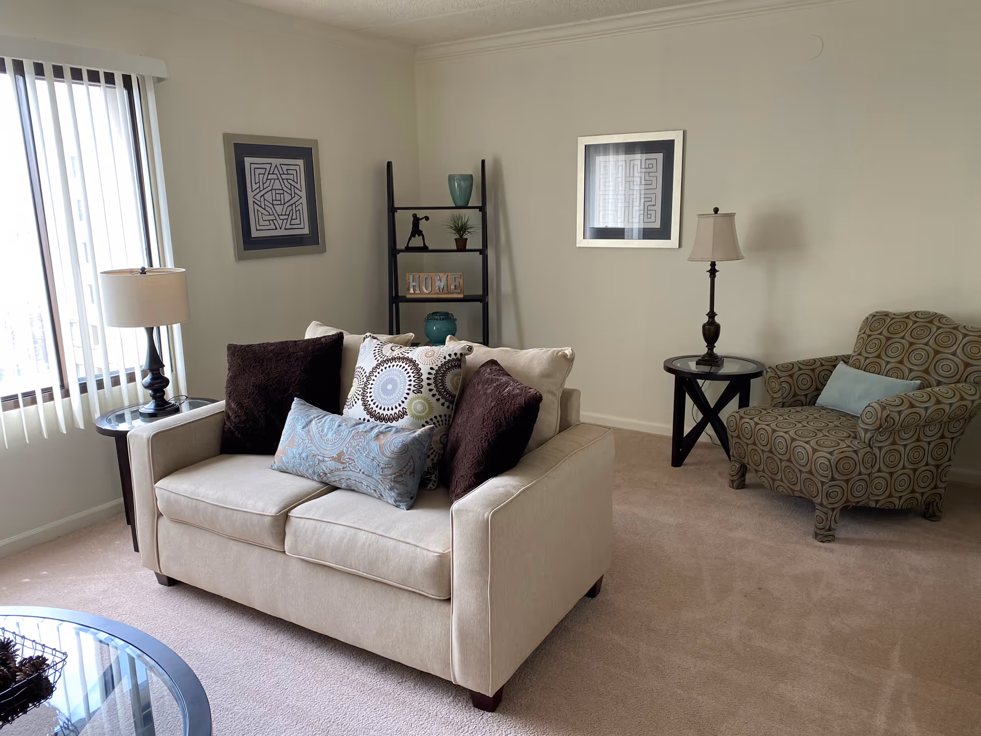 A bright living room with a beige loveseat topped with patterned pillows, a patterned armchair, side tables with lamps, a decorative shelf and framed wall art.