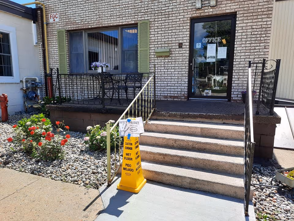 Entrance to a building with a small set of concrete stairs leading up to a door labeled 'OFFICE'. There is a yellow caution sign on the sidewalk warning of a wet floor and wet paint. To the left of the stairs is a small garden area with red and pink flowers and a black metal patio table with chairs on a raised platform. The building exterior is made of light-colored brick with green shutters on the window.