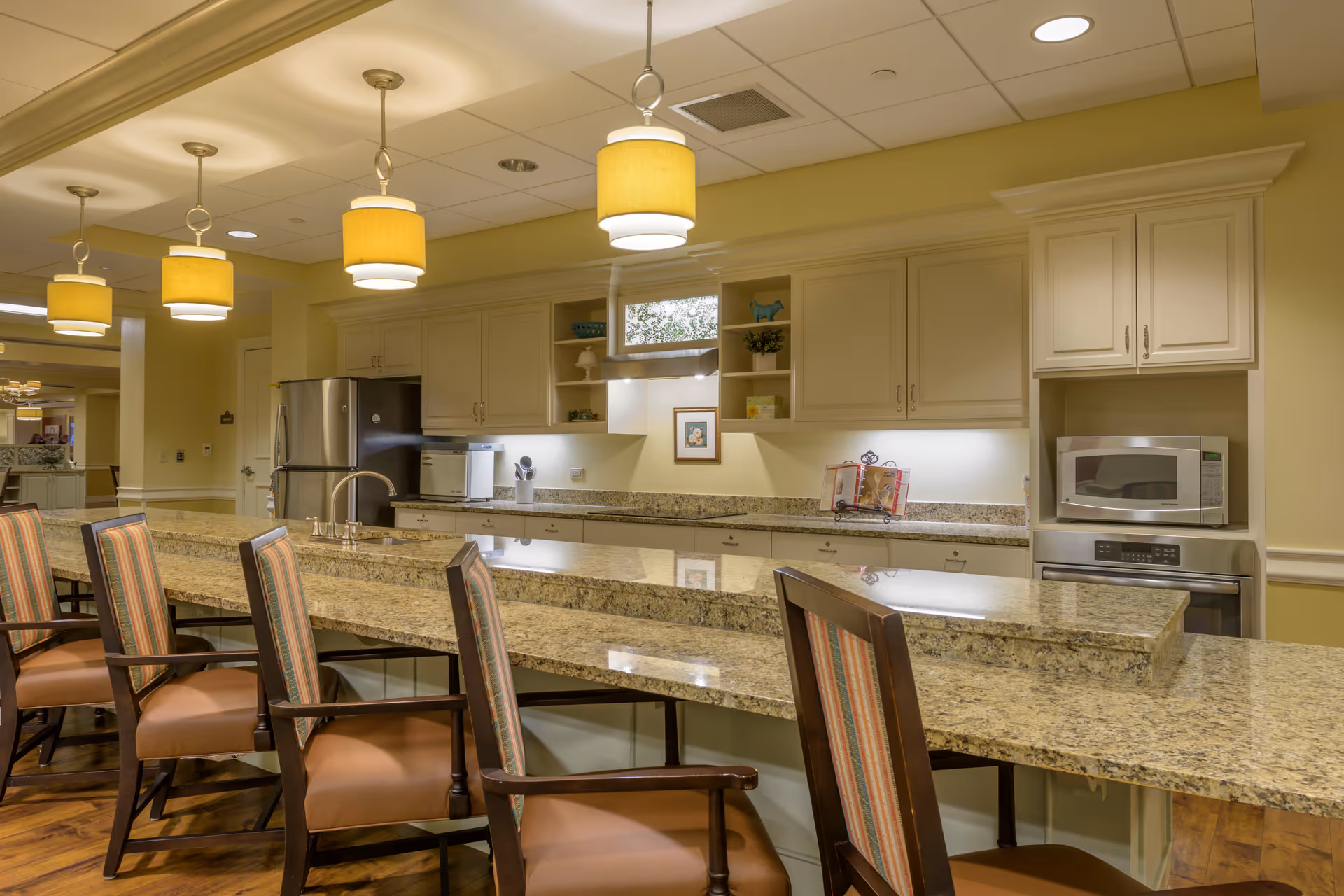 A well-lit kitchen area with granite countertops and several high-backed chairs with striped upholstery. The kitchen features cream-colored cabinets, a stainless steel refrigerator, a microwave, an oven, and pendant lights hanging from the ceiling. The space has a warm and inviting atmosphere.