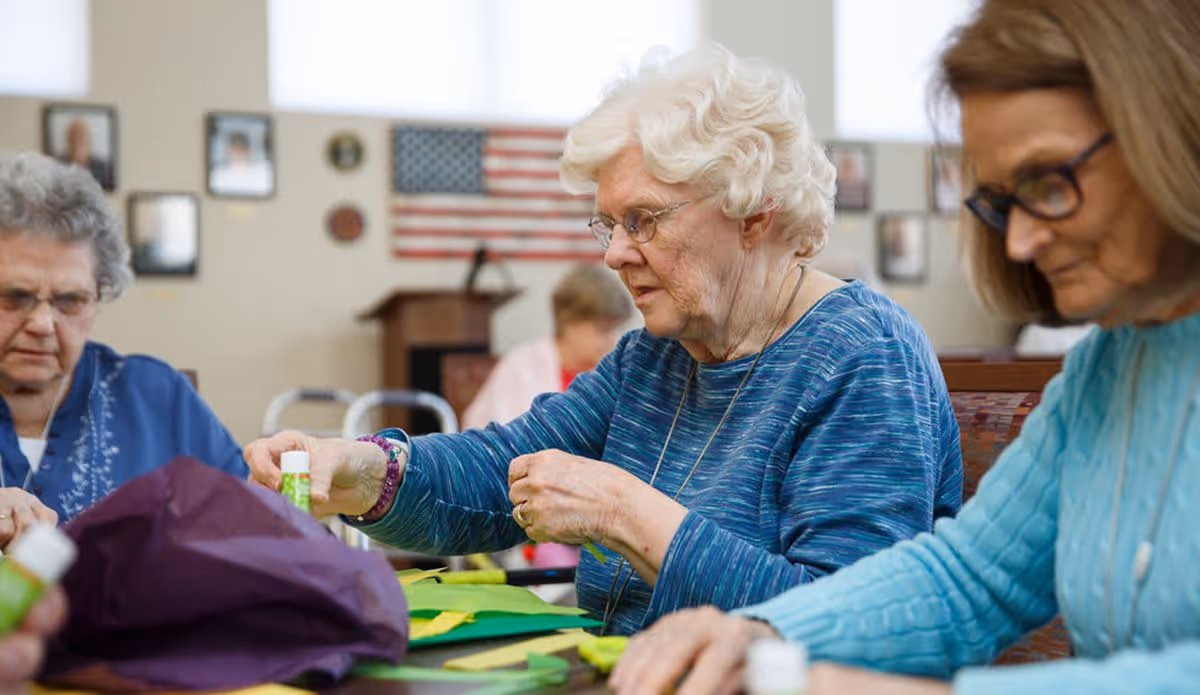 Three elderly women sitting at a table engaged in a craft activity, using glue sticks and colored paper, with an American flag and framed pictures on the wall in the background.