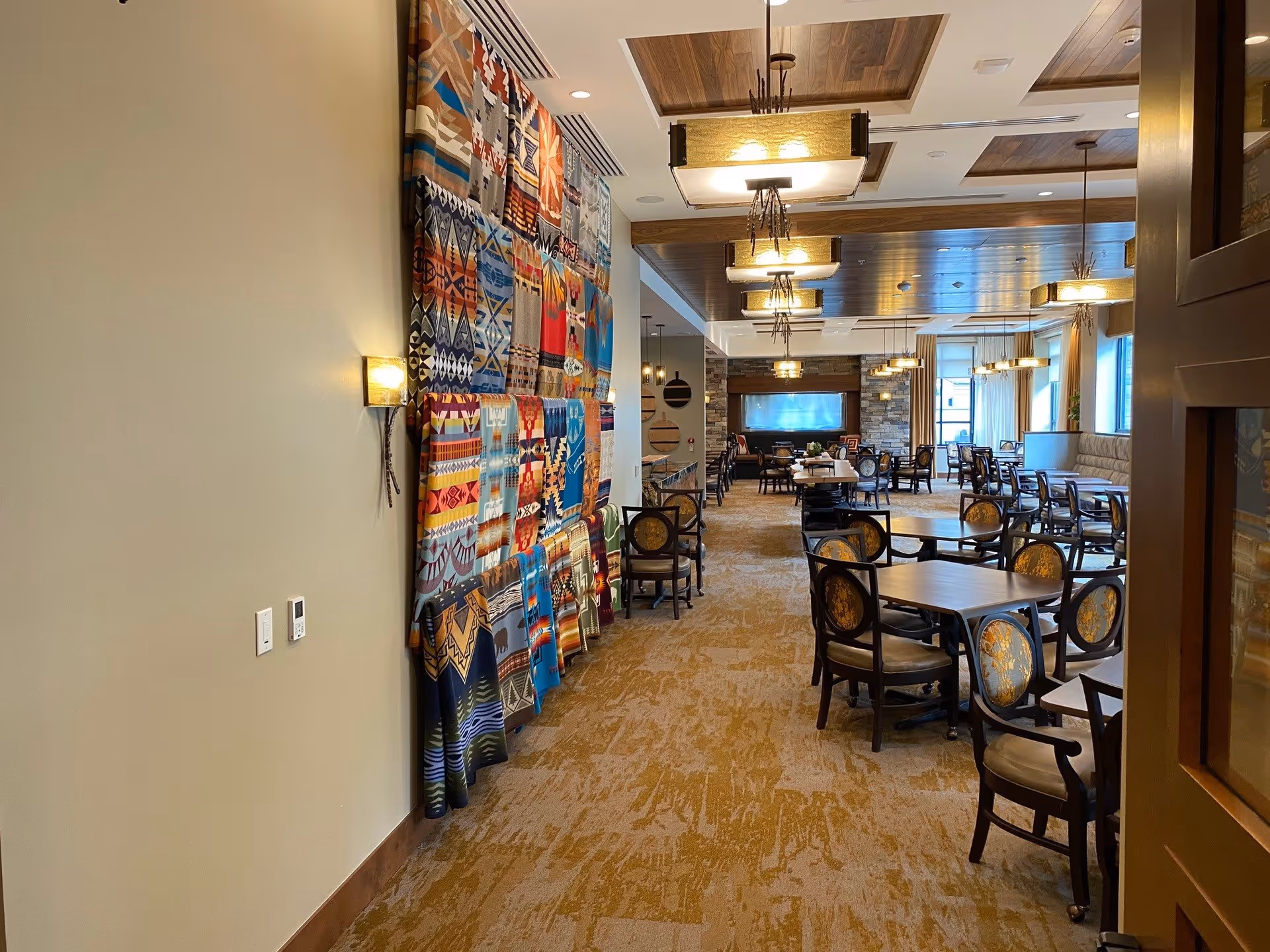 Interior view of a dining room in a senior living facility with multiple round tables and chairs arranged neatly. The room features a patterned carpet, wooden ceiling panels with hanging light fixtures, and a large colorful textile art piece on the left wall. Large windows in the background allow natural light to brighten the space.