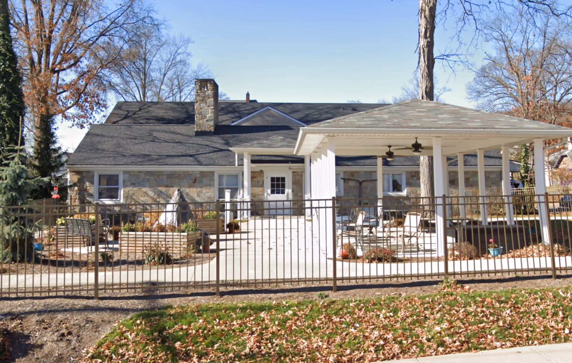 Exterior view of a single-story stone building with a dark shingled roof and a chimney. In front of the building is a fenced patio area with raised garden beds, outdoor seating, and a covered pavilion with ceiling fans. Trees with bare branches surround the area, and fallen leaves cover the ground.