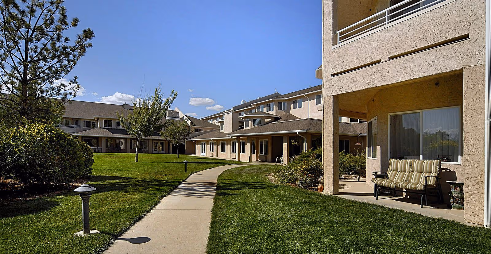 Outdoor view of a senior living facility with a paved walkway surrounded by green grass, bushes, and trees. The building has beige walls with multiple windows and a covered patio area with cushioned seating. The sky is clear and blue with a few clouds.
