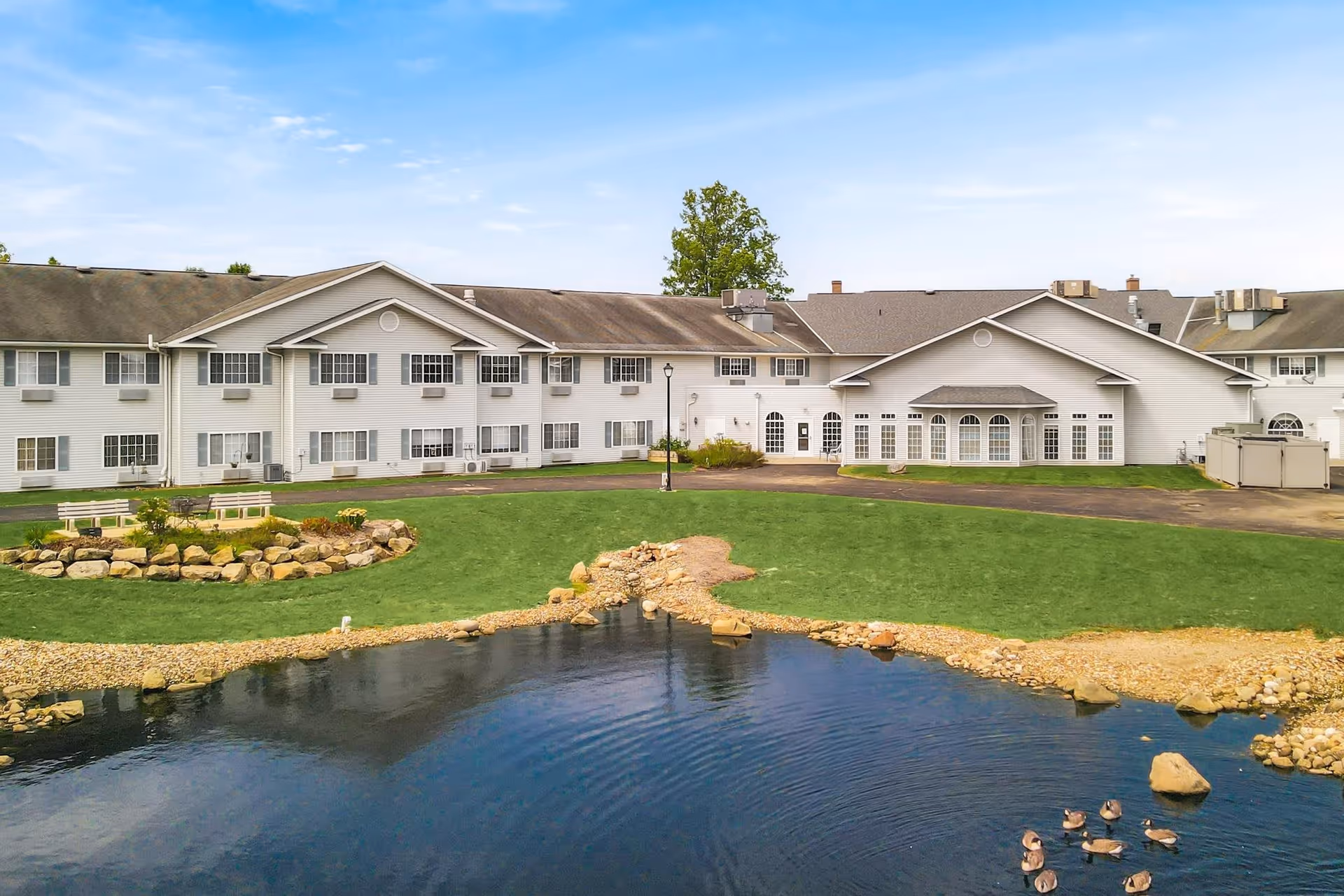 Exterior view of a two-story senior living facility building with white siding, multiple windows, and a well-maintained lawn. In the foreground, there is a small pond with ducks swimming and a landscaped area with rocks and benches. The sky is clear with a few clouds.
