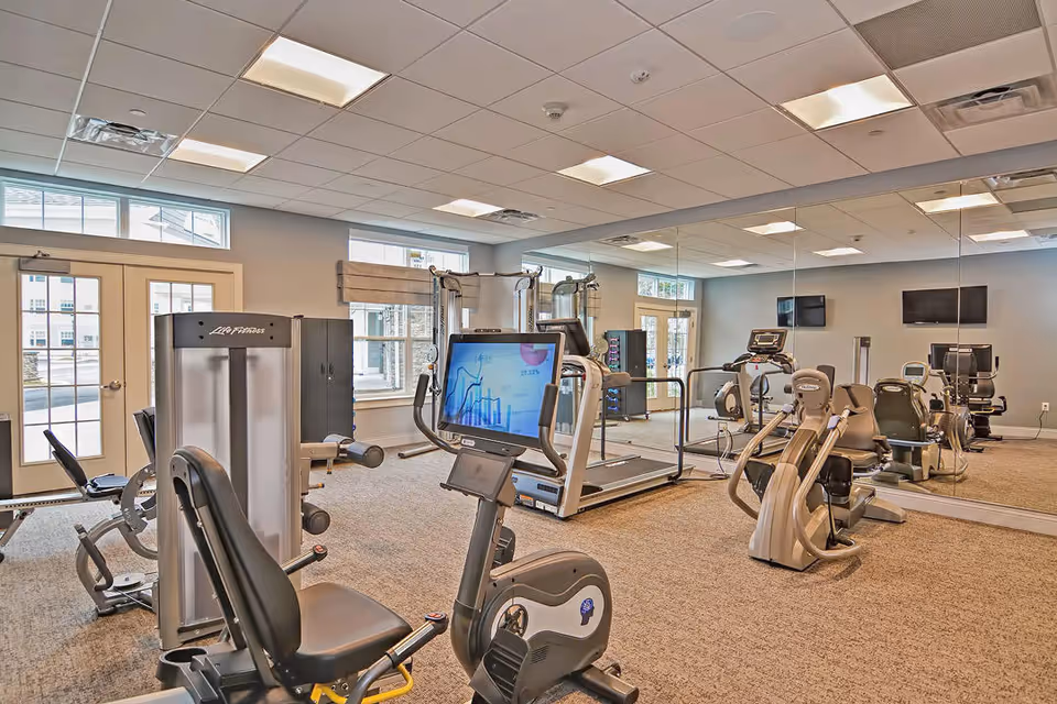 A well-lit fitness room in a retirement community featuring various exercise equipment including stationary bikes, treadmills, and weight machines. The room has large windows, mirrored walls, and a carpeted floor.