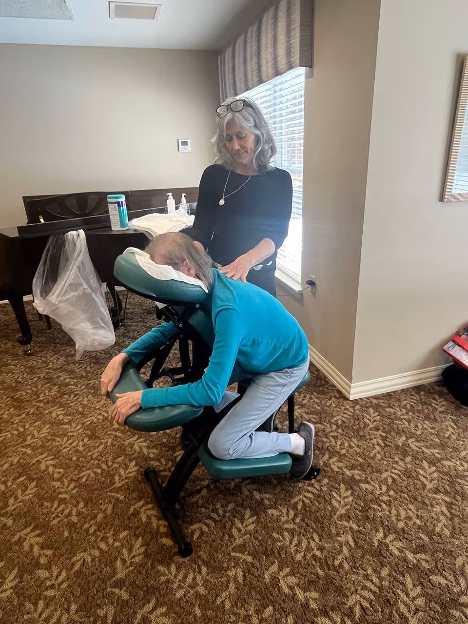 A woman with gray hair wearing a black top is giving a seated massage to an elderly person in a teal sweater who is resting on a green massage chair in a carpeted room with a window and a piano in the background.
