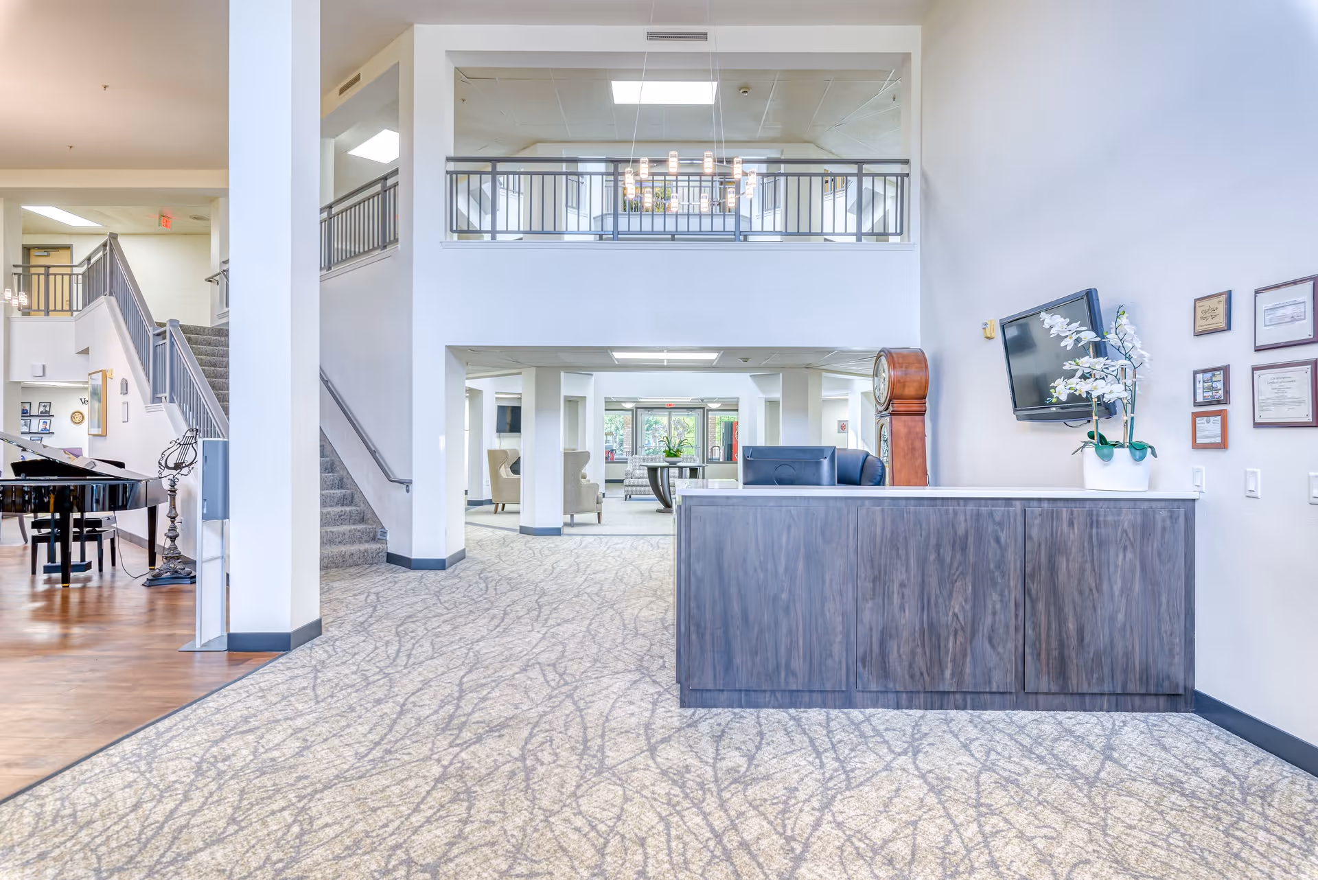 Bright and spacious senior living facility lobby with a reception desk on the right, a large grandfather clock behind it, and a flat-screen TV mounted on the wall. The area features carpeted flooring with a patterned design, white walls, and a staircase leading to an upper floor. To the left, there is a grand piano on a wooden floor section, and comfortable seating areas are visible in the background.