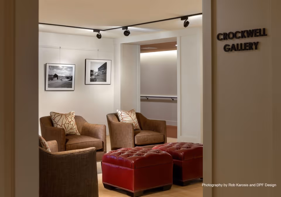 Interior view of a gallery lounge area named Crockwell Gallery with three brown armchairs each with a decorative pillow, two red tufted ottomans, and two black and white framed photographs on the wall.