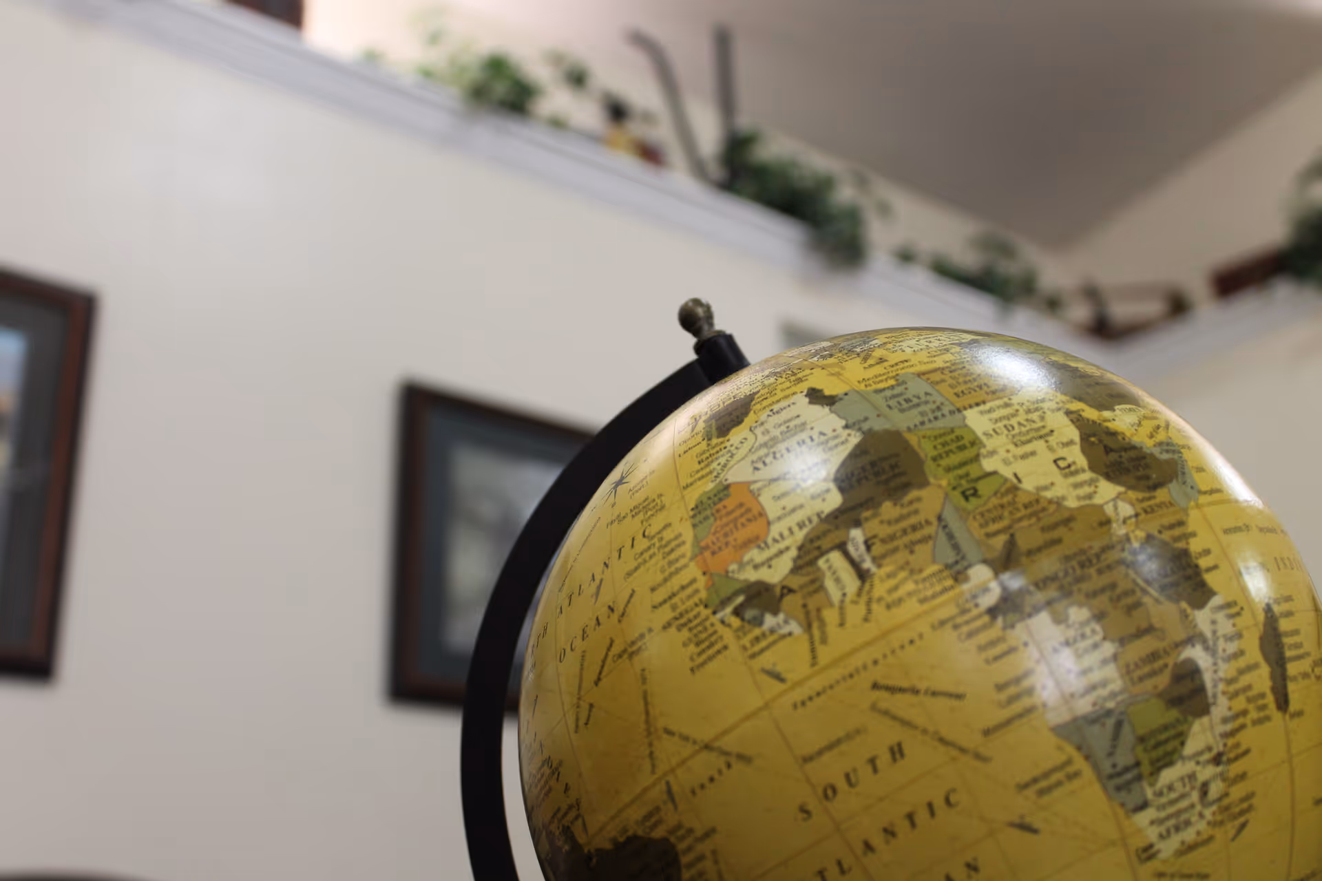 Close-up view of a yellow globe showing parts of Europe and Africa, with a blurred background featuring a white wall, two framed pictures, and a shelf with green plants.