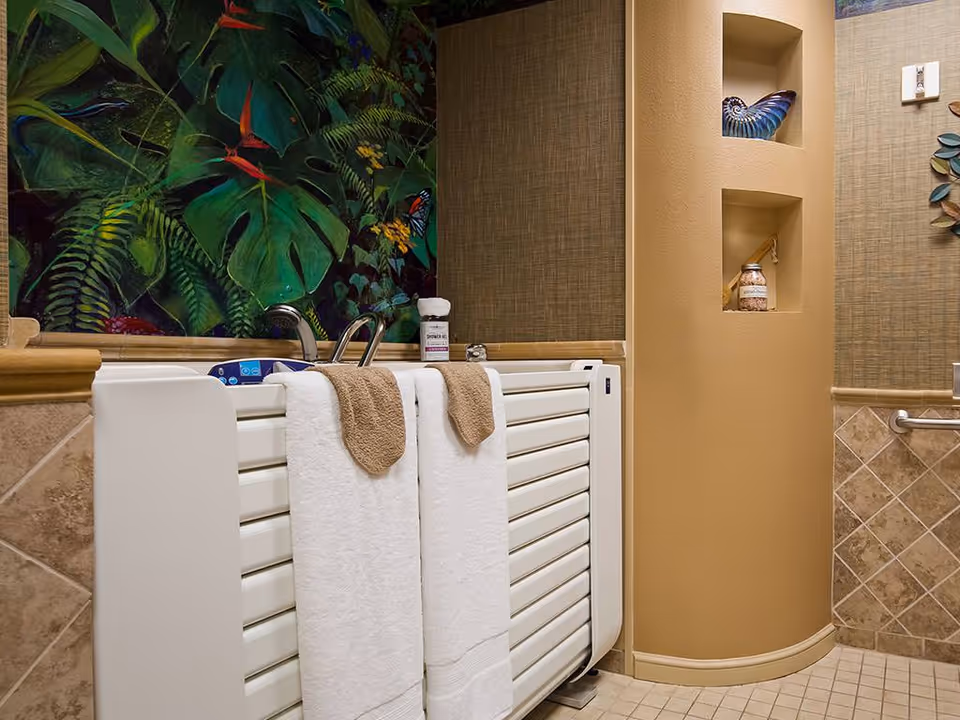 A bathroom featuring a white walk-in bathtub with two white towels hanging on the side, beige tiled walls and floor, a decorative tropical-themed wall art above the tub, and built-in wall niches holding decorative items.