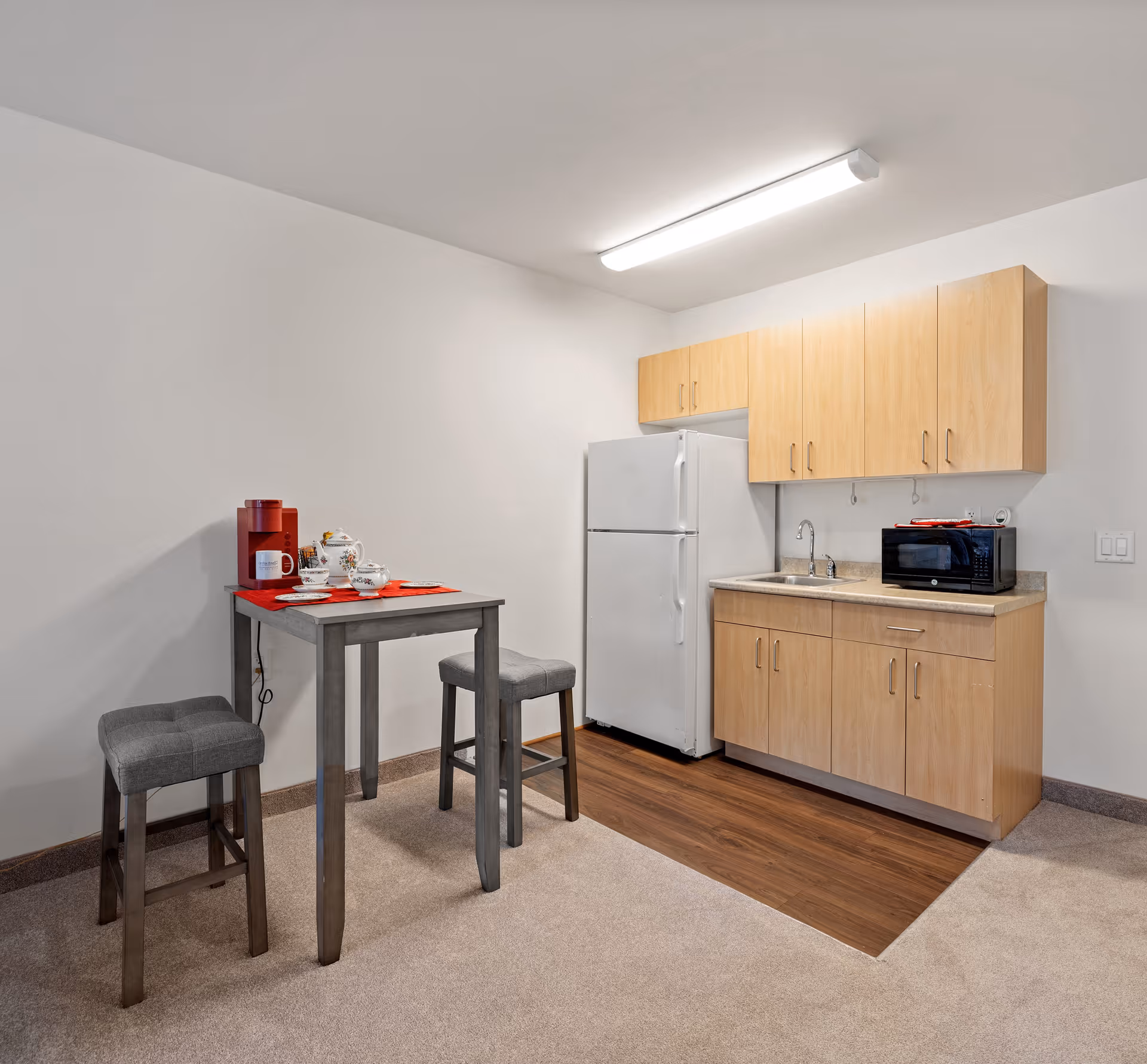 Small kitchenette area with light wood cabinets, a white refrigerator, a microwave on the countertop, and a sink. Adjacent to the kitchenette is a small dining table with two gray cushioned stools. The table has a red placemat, a red coffee maker, a white mug, and a tea set. The floor is a combination of carpet and wood laminate.